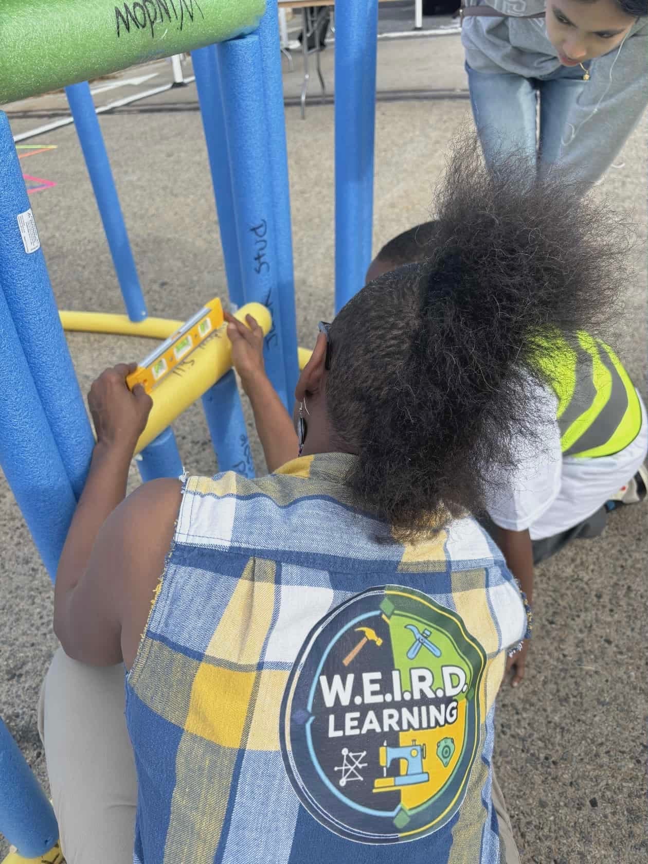 kito morris kneeling on the street, W.E.I.R.D. learning logo visible on the back of her denim vest, helping a kid level a frame