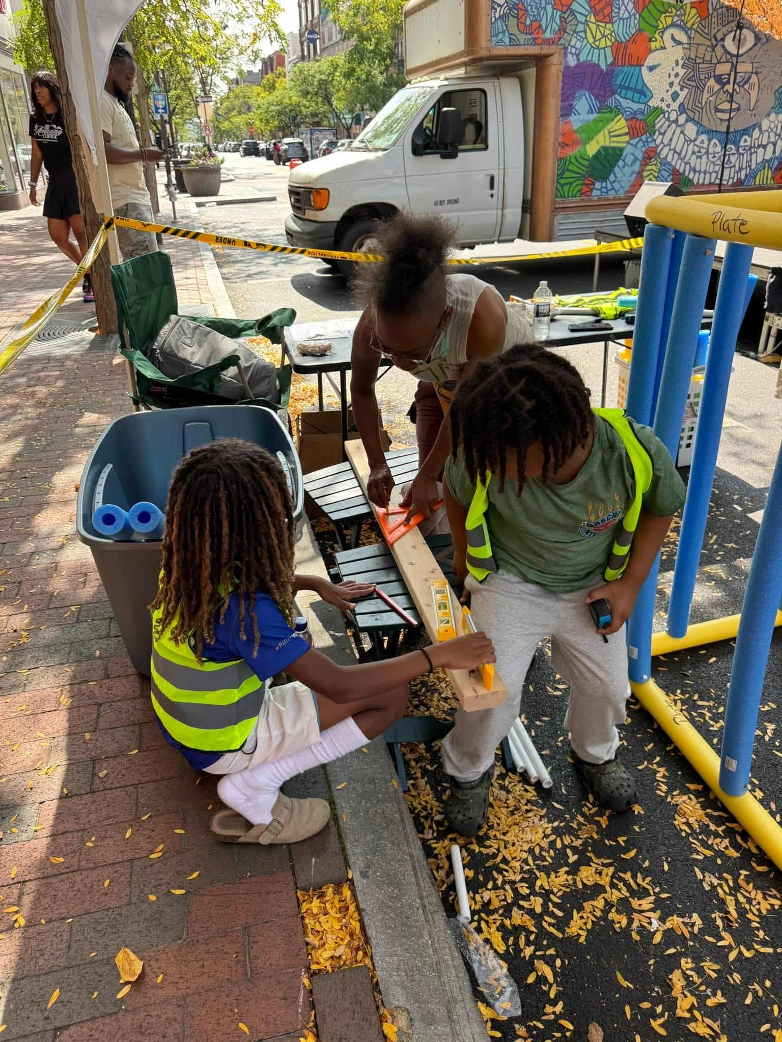 kids in safety vests building a pool-noodle house frame on the street during a community festival, surrounded by neighbors and music